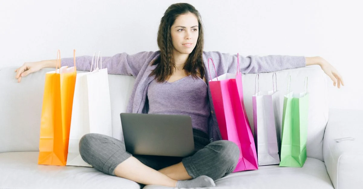 Jeune femme assise sur un canapé avec des sacs de shopping colorés et un ordinateur portable, représentant le shopping en ligne.