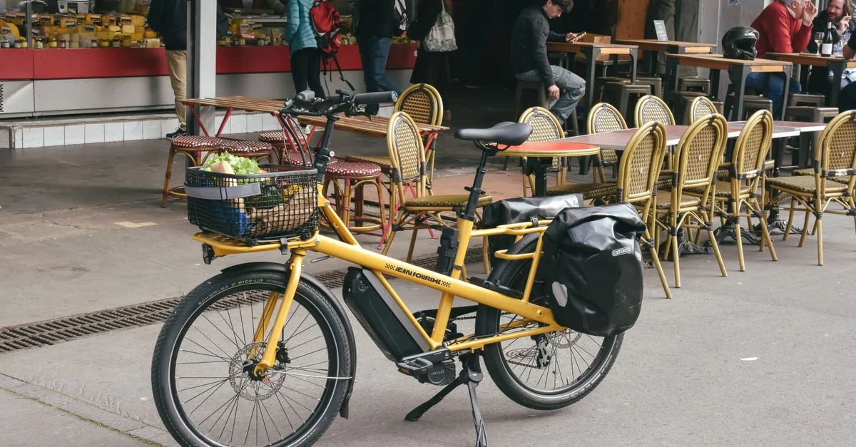Vélo cargo jaune garé dans une rue devant un café en France, ajoutant une scène de rue animée.